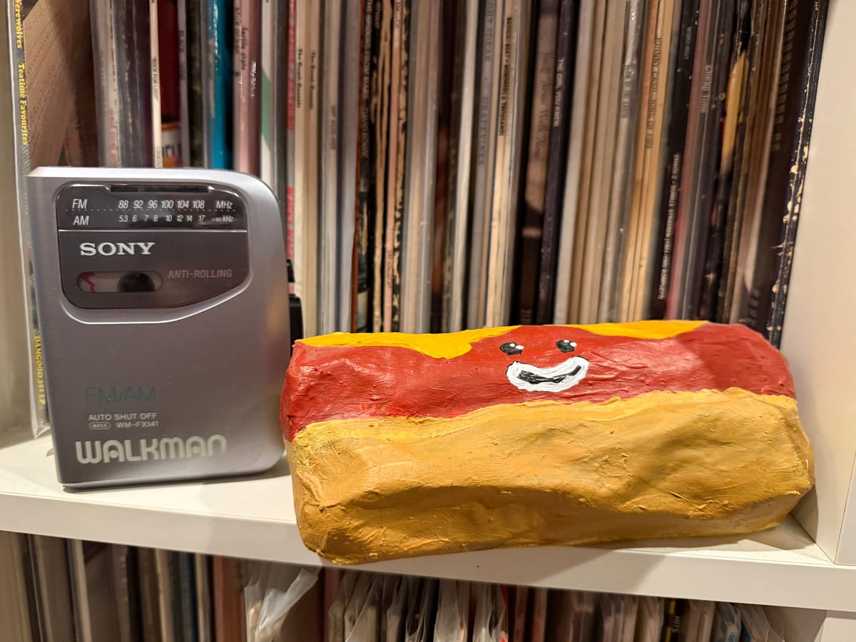 A silver Sony Walkman with a radio standing on a shelf with a paper mache hot dog next to it. The hotdog has a smiling face. There are 12" vinyl records on shelved behind them.