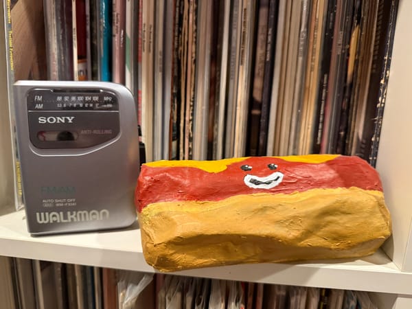 A silver Sony Walkman with a radio standing on a shelf with a paper mache hot dog next to it. The hotdog has a smiling face. There are 12" vinyl records on shelved behind them.